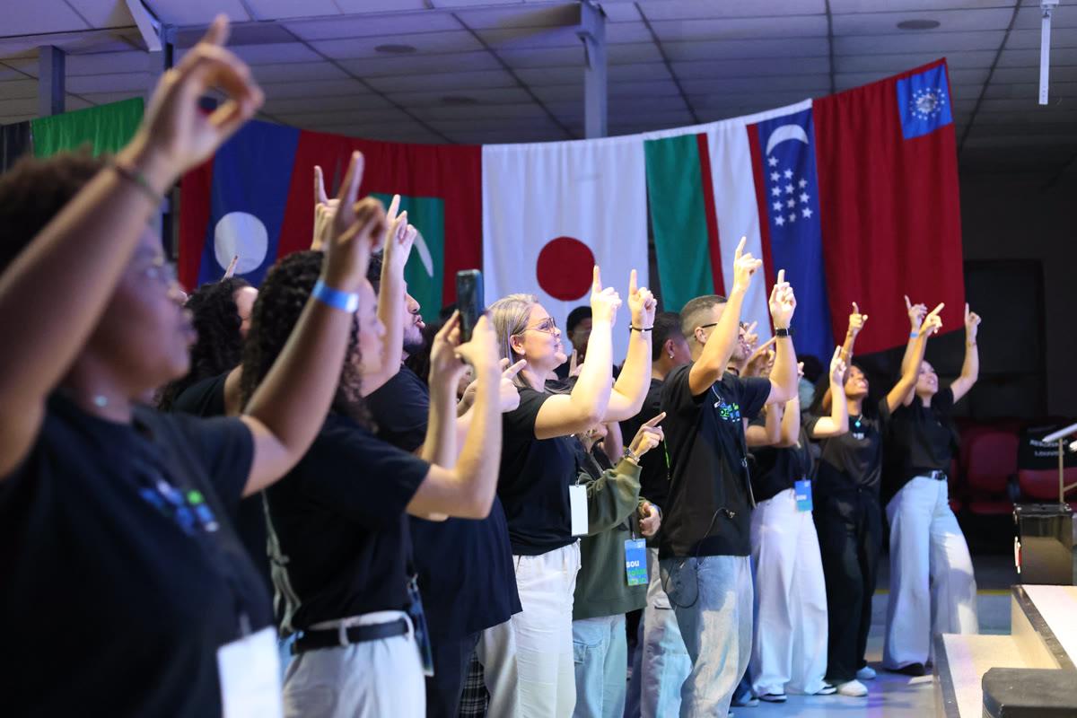 Pessoas de braços erguidos, durante um workshop de missões, diante de bandeiras de várias nações